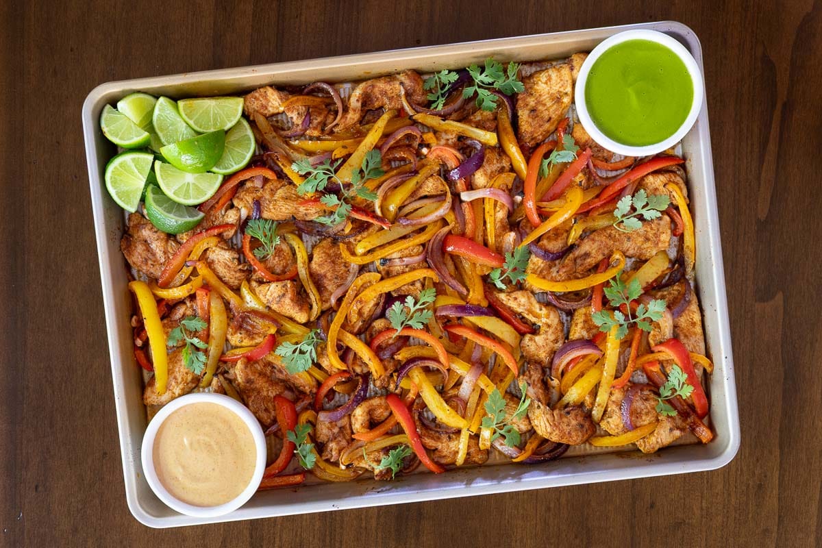 Overhead photo of Sheet Pan Chicken Fajitason a wood table with bowls of Mexican Cilantro Sauce and Chipotle Sauce with lime wedges.