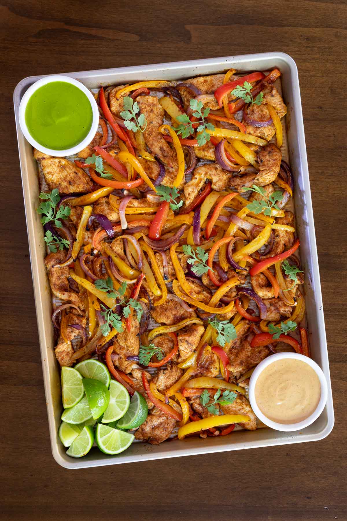 Overhead photo of Sheet Pan Chicken Fajitason a wood table with bowls of Mexican Cilantro Sauce and Chipotle Sauce with lime wedges.