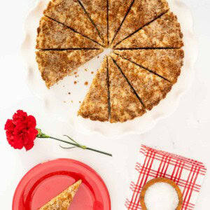 Overhead view of a round Biscoff shortbread sliced into wedges on a white scalloped plate, with one slice served on a red plate beside a red carnation and flaked sea salt.