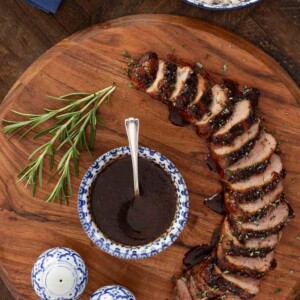 Sliced grilled pork tenderloin with balsamic fig glaze on a wooden board, served with a bowl of glaze, a sprig of rosemary, and a side of white rice in a blue and white bowl.