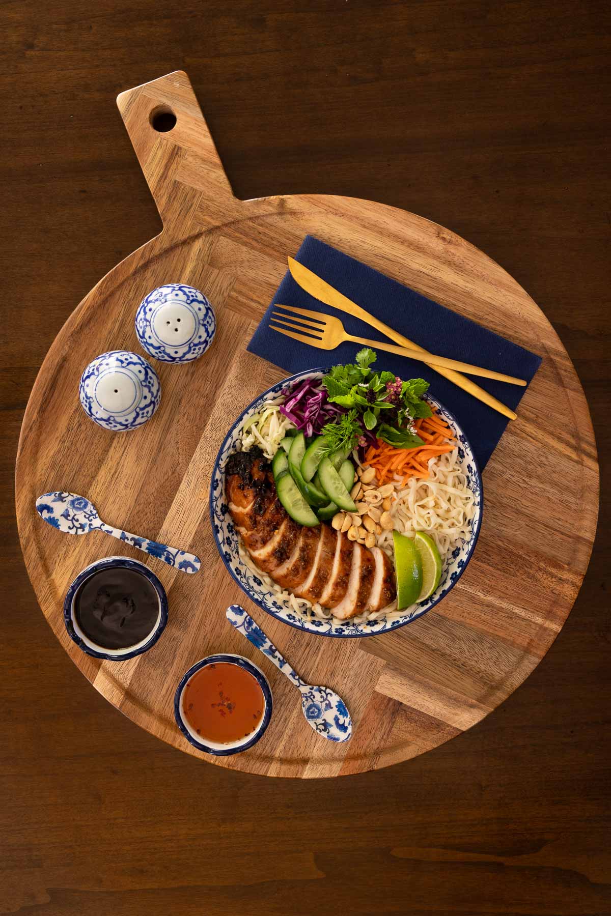 Overhead view of a finished lemongrass grilled chicken bowl with rice noodles, colorful vegetables, peanuts, and herbs, served with hoisin and nuoc cham sauces on a wooden board alongside gold utensils and blue-and-white dishes.
