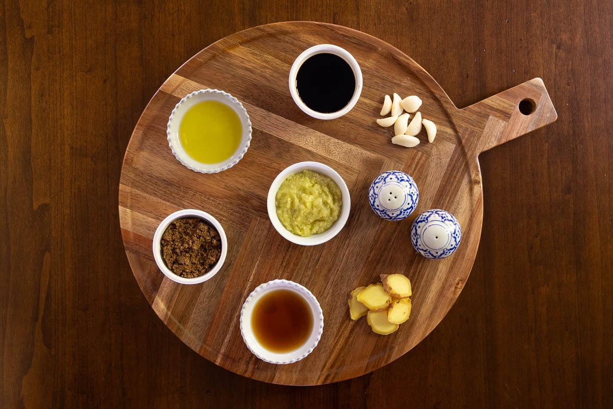 Overhead view of lemongrass chicken marinade ingredients arranged on a wooden round board, including brown sugar, soy sauce, oil, garlic, fresh ginger, fish sauce, and a small bowl of lemongrass paste.