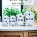 Four white and blue ceramic canisters labeled “The Café Sucre Farine” sitting on a kitchen counter in front of a sunny window with fresh plants in the background.
