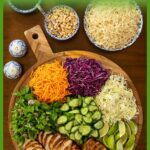 Overhead view of sliced lemongrass grilled chicken on a round wooden board surrounded by fresh herbs, shredded carrots, red and green cabbage, cucumbers, lime wedges, and garnishes, with bowls of noodles, peanuts, hoisin sauce, and nuoc cham on the side.