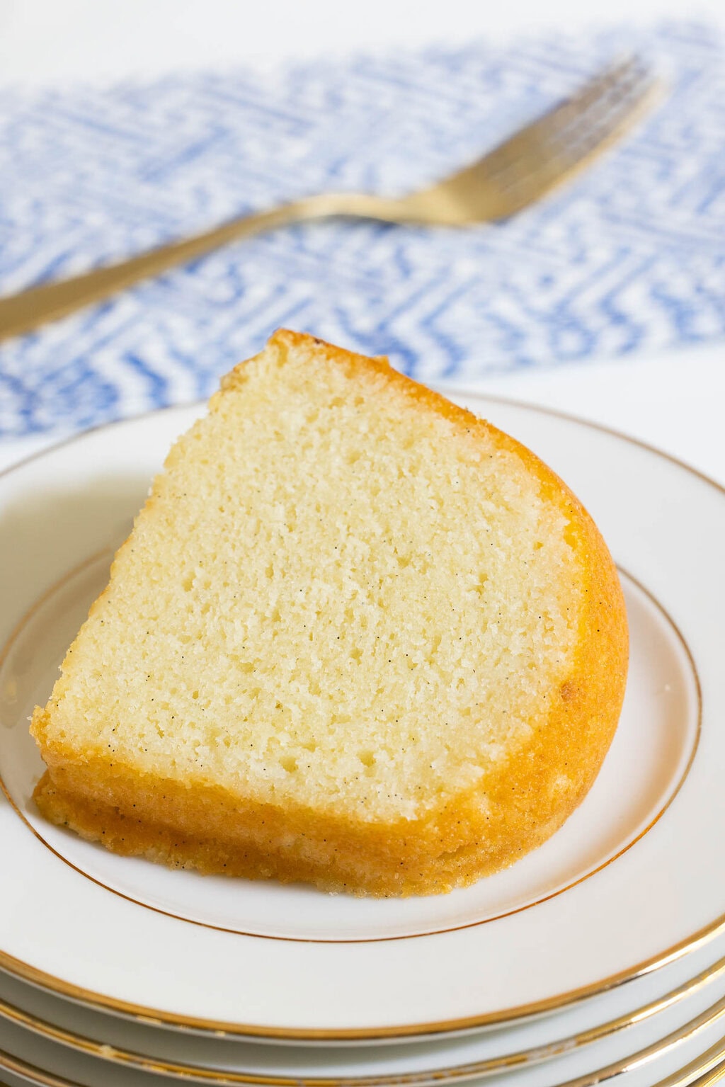 Slice of Vanilla Bean Bundt Cake on a stack of white china plates with gold trim, showing the moist, tender crumb flecked with vanilla bean
