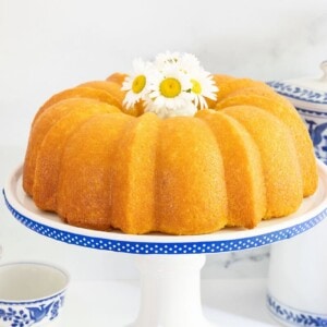Golden Vanilla Bean Bundt Cake with a shiny glaze, displayed on a white cake stand with blue polka dots, topped with a small vase of fresh daisies. Blue and white china cups and a matching teapot in the background.