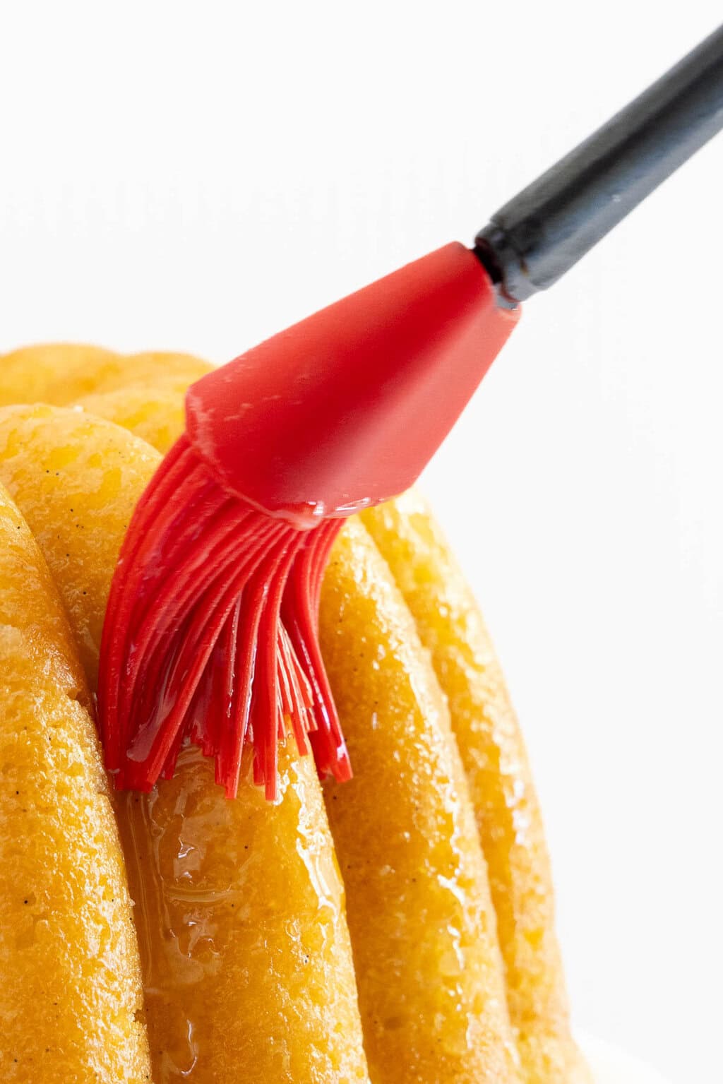 close up of the glaze being applied with a red silicon pastry brush onto a golden vanilla bean bundt cake