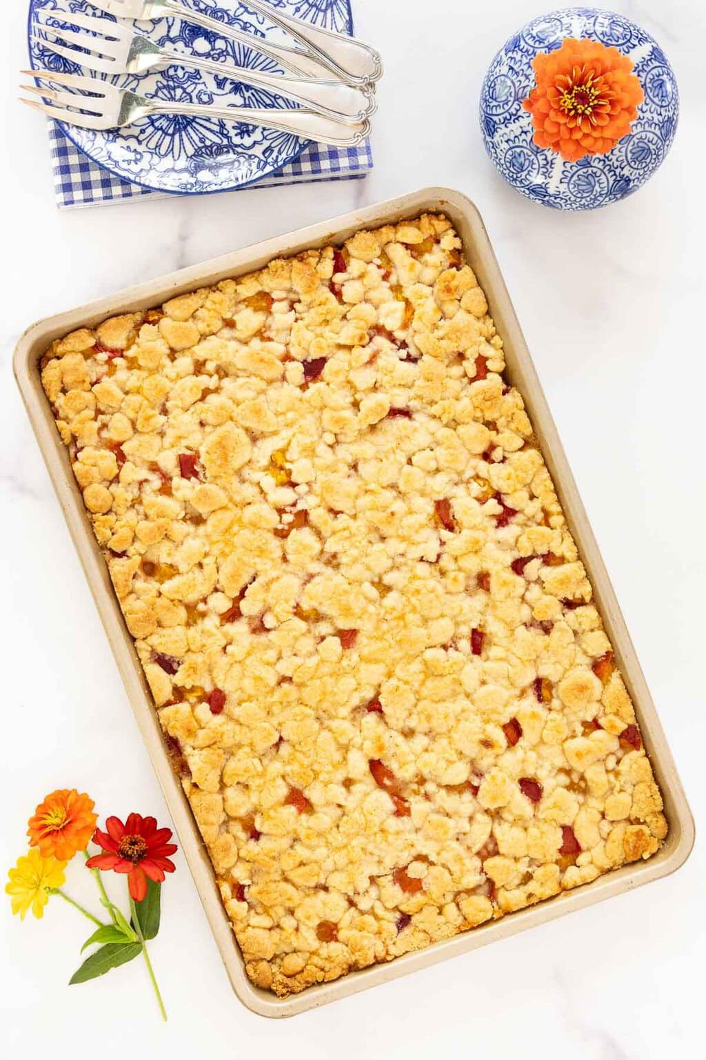 Golden peach crumb bars in a pan,  next to blue floral serving plates, forks with fresh zinnias, a perfect summer dessert.