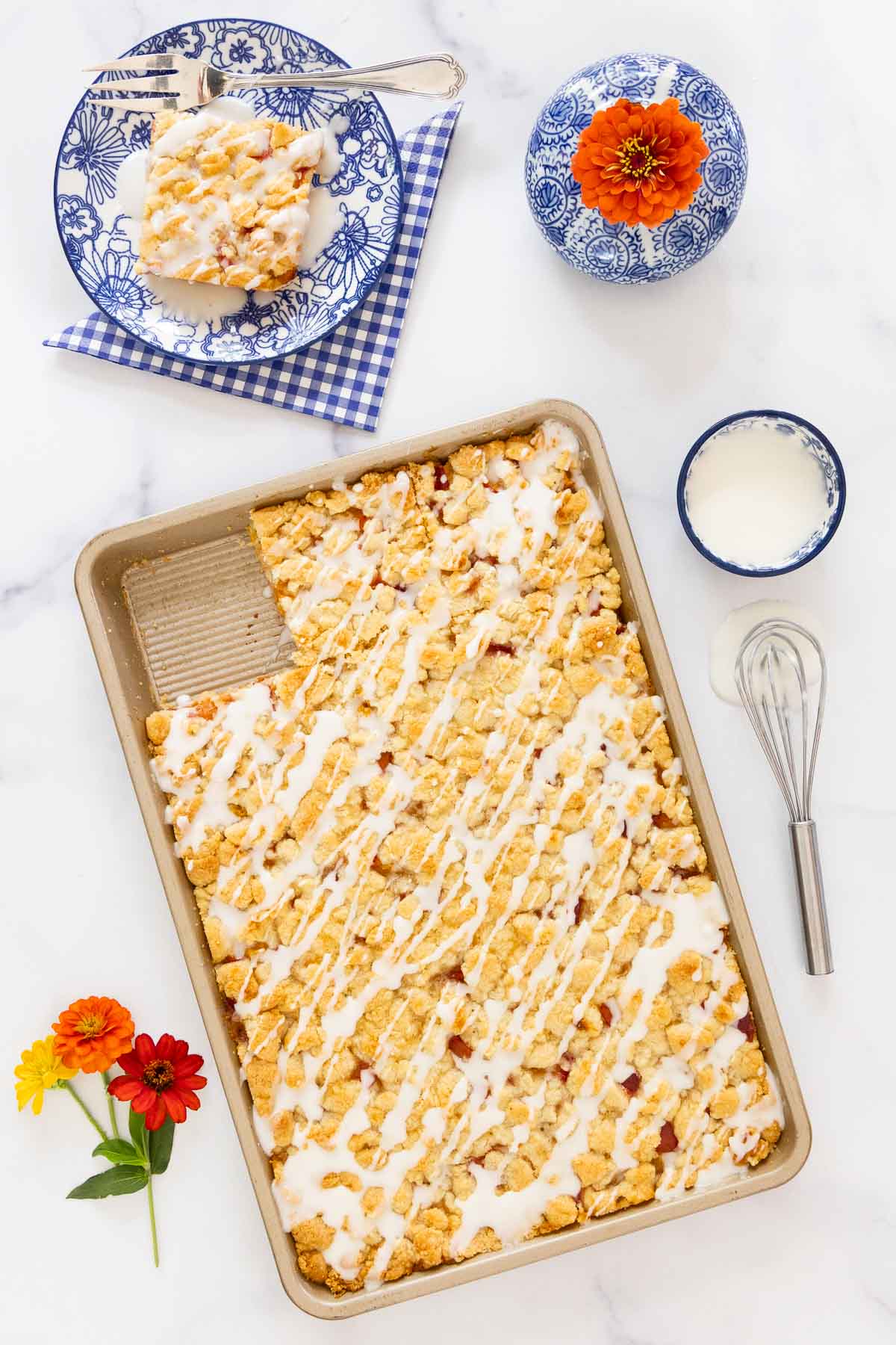 Full pan of peach crumb bars drizzled with almond glaze, one square removed and placed on an individual blue-patterned serving plate with a whisk and icing bowl nearby.