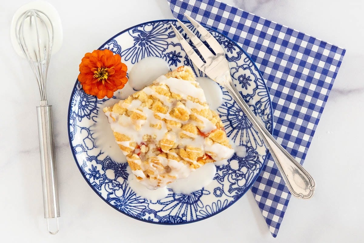 Iced peach crumb bar served on a blue floral plate with almond drizzle and a small whisk.