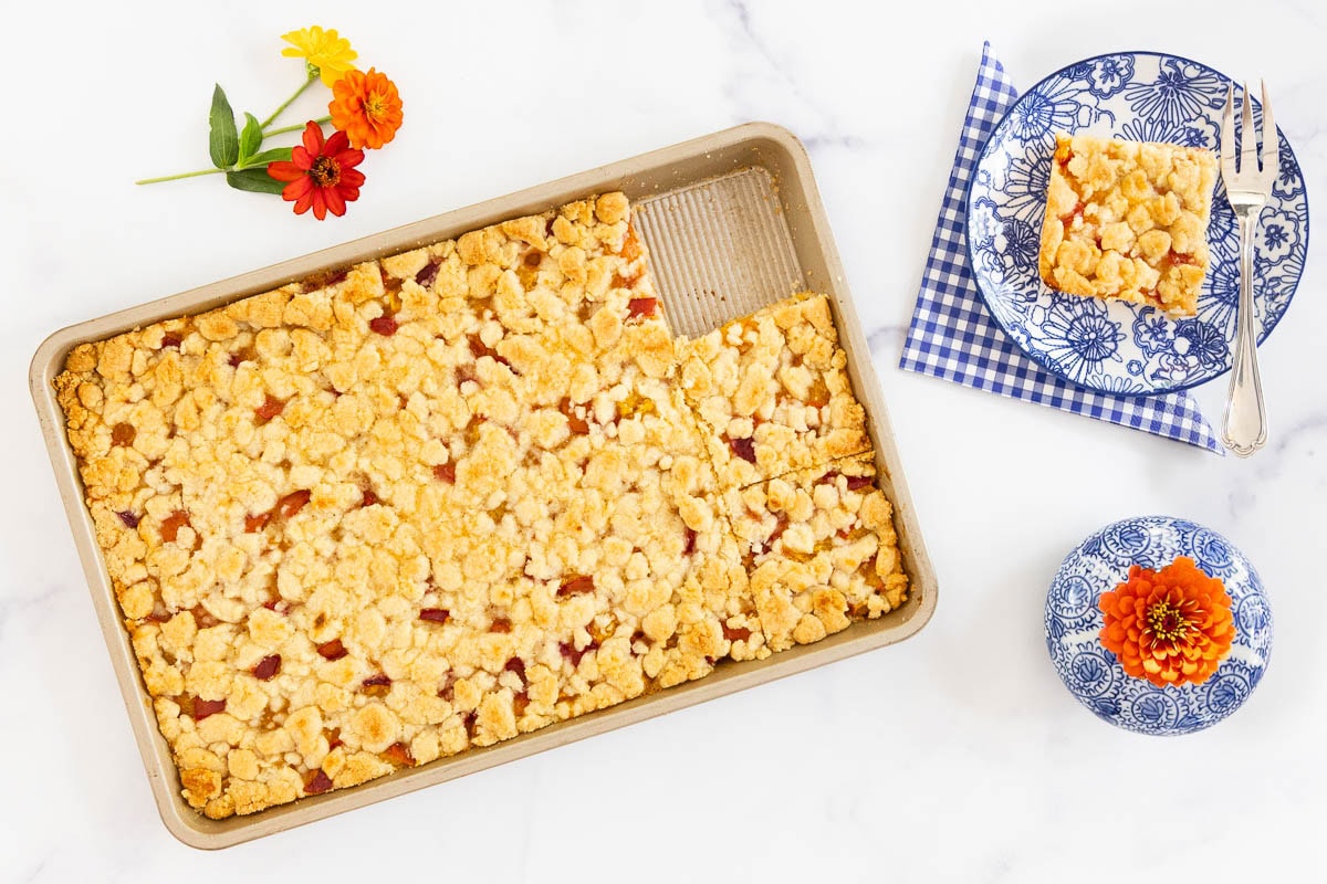 Pan of buttery peach crumb bars with one square served on a floral plate.