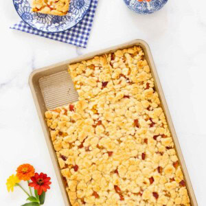 Peach crumb bars in a pan with one square served on a blue floral plate, garnished with fresh flowers.