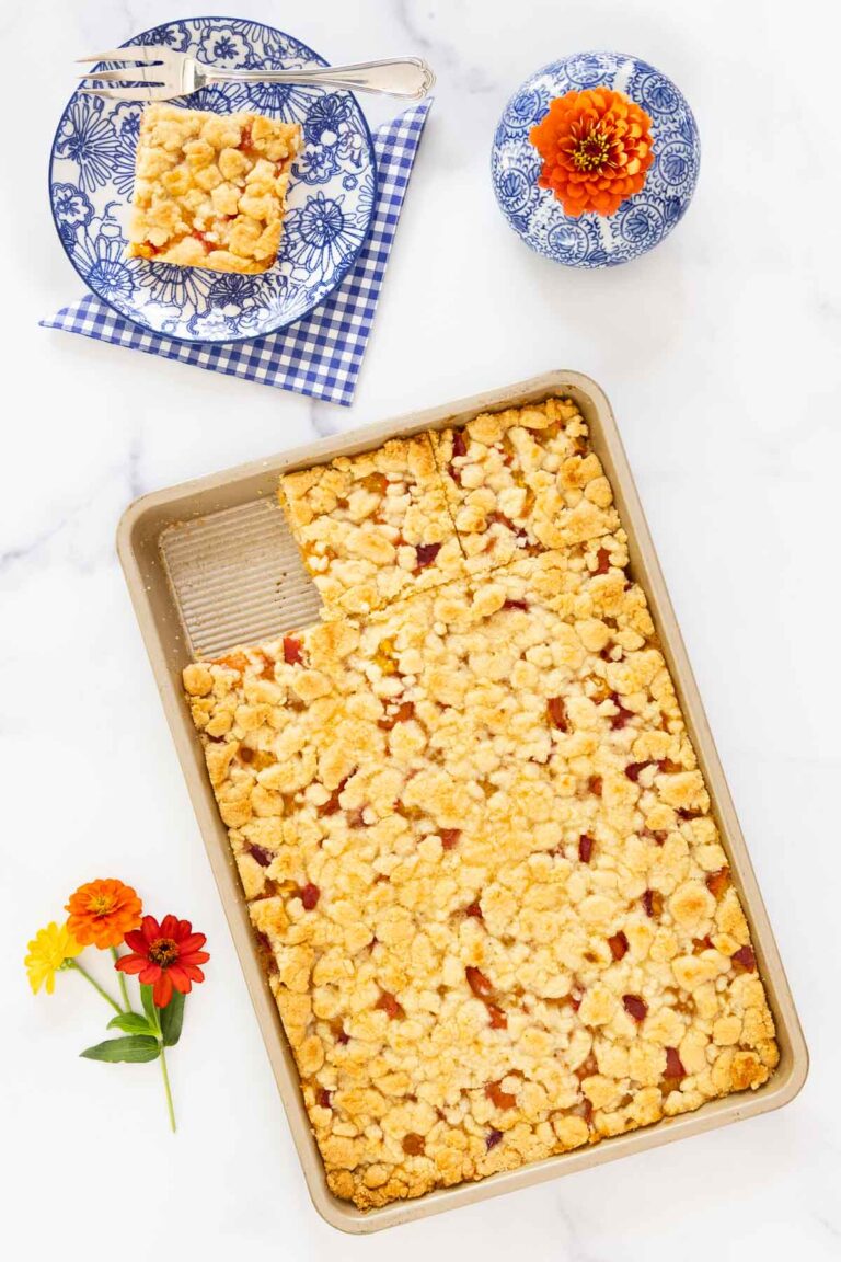Peach crumb bars in a pan with one square served on a blue floral plate, garnished with fresh flowers.