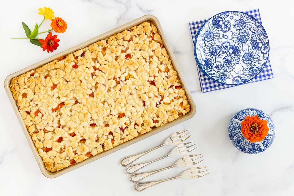 Golden peach crumb bars in a pan,  blue floral plates with fresh zinnias, a perfect summer dessert.