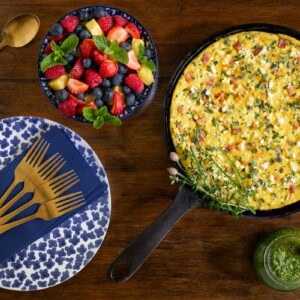 Overhead view of a ham and cheese frittata with goat cheese and fresh herbs in a cast iron skillet, served with a bowl of fresh fruit, a jar of green herb sauce, and a blue floral plate with gold forks-perfect for a make-ahead brunch.