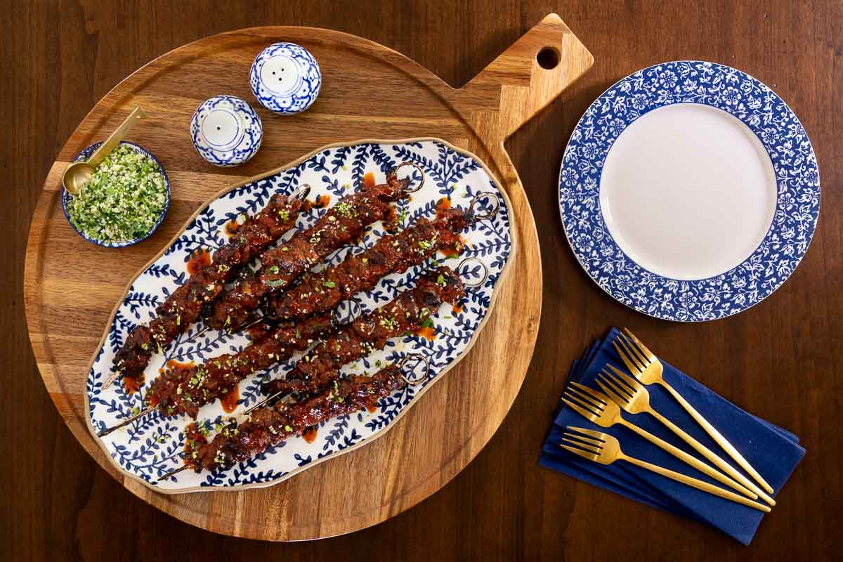 Grilled Korean beef skewers on a patterned platter with gremolata, salt and pepper shakers, and styled tableware.