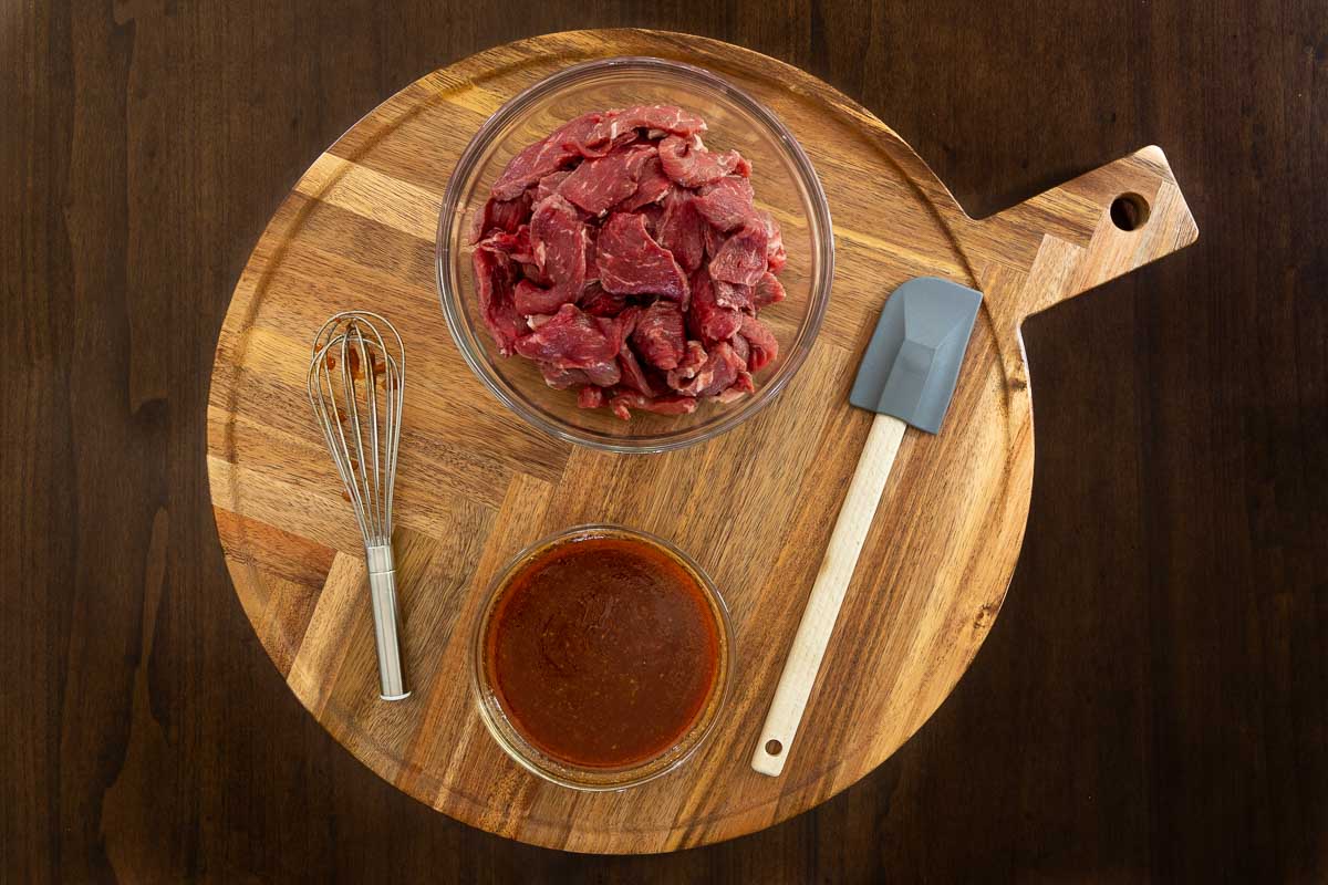 Sliced raw beef in a glass bowl next to a whisked Korean-style marinade with tools ready for mixing on a wooden board