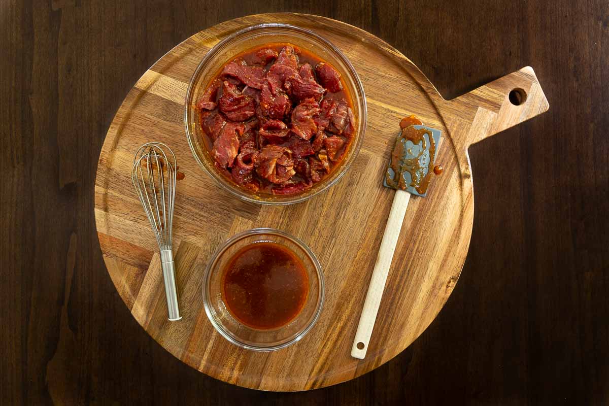 Glass bowls of sliced raw beef and Korean-style marinade on a round wooden board with whisk and spatula.