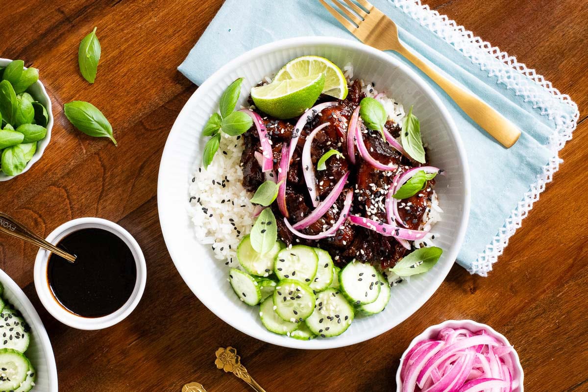 A honey soy rice bowl with glazed meat, pickled red onions, cucumber slices, sesame seeds, fresh basil, and lime wedges on a wooden table.