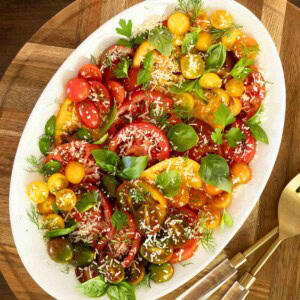 Heirloom and cherry tomato salad with fresh herbs and grated Parmesan on a white platter, served on a wooden board.