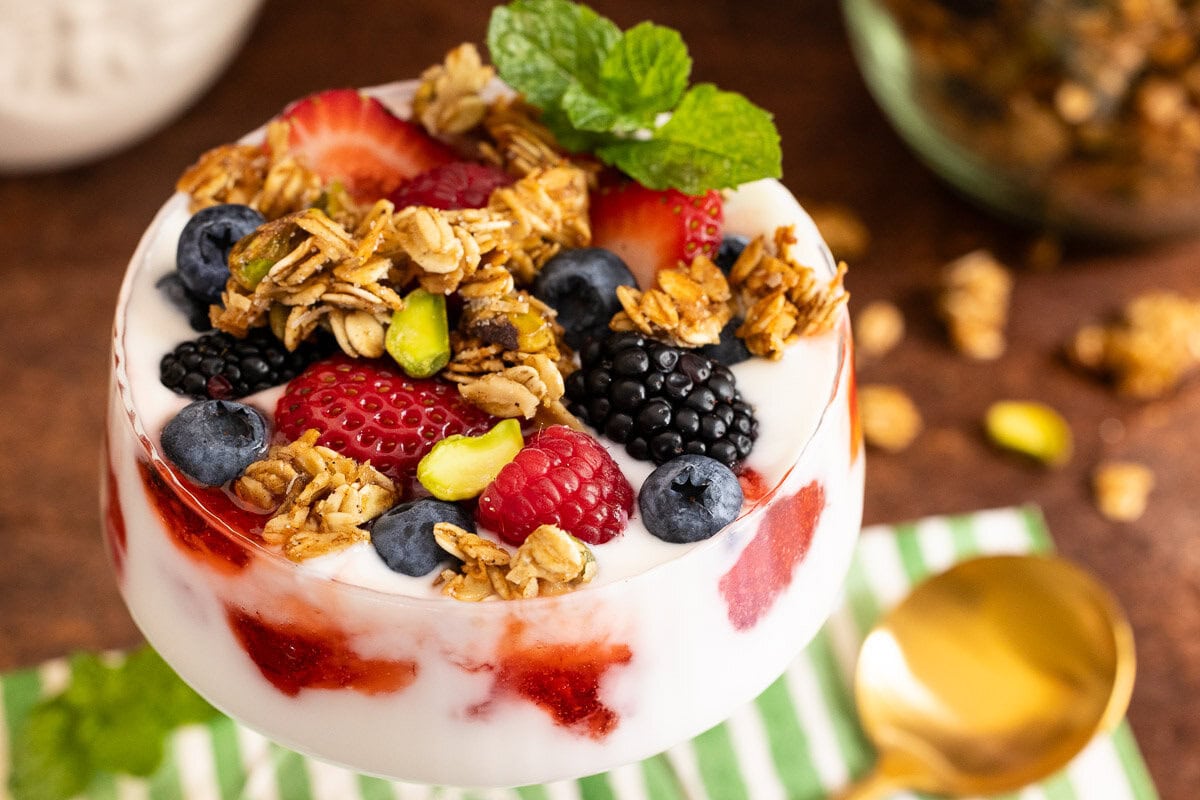 Glass filled with yogurt, fresh berries, and crunchy Maple Pistachio Granola topped with mint, served with a gold spoon.