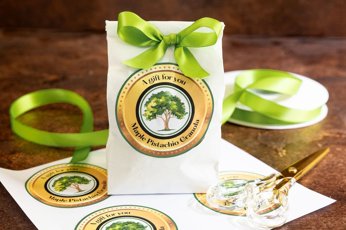 Bag of homemade Maple Pistachio Granola decorated with a printable gift label and green ribbon, surrounded by extra labels, ribbon, and scissors.