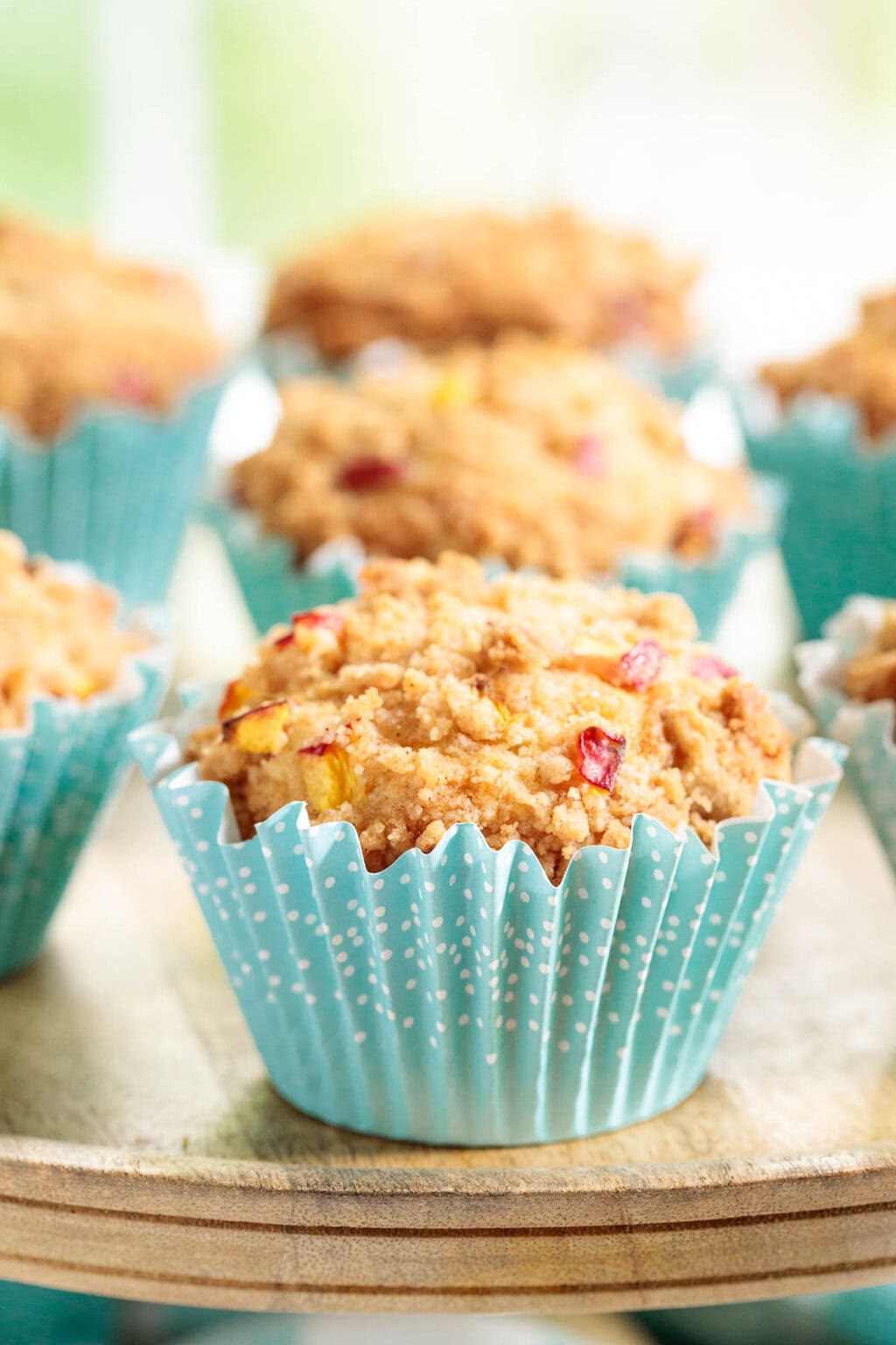 Homemade peach crumble muffins with fresh peaches and buttery streusel topping, served in blue polka dot cupcake wrappers on a wooden cake stand.