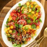 Heirloom and cherry tomato salad with fresh herbs and grated Parmesan on a white platter, served on a wooden board.