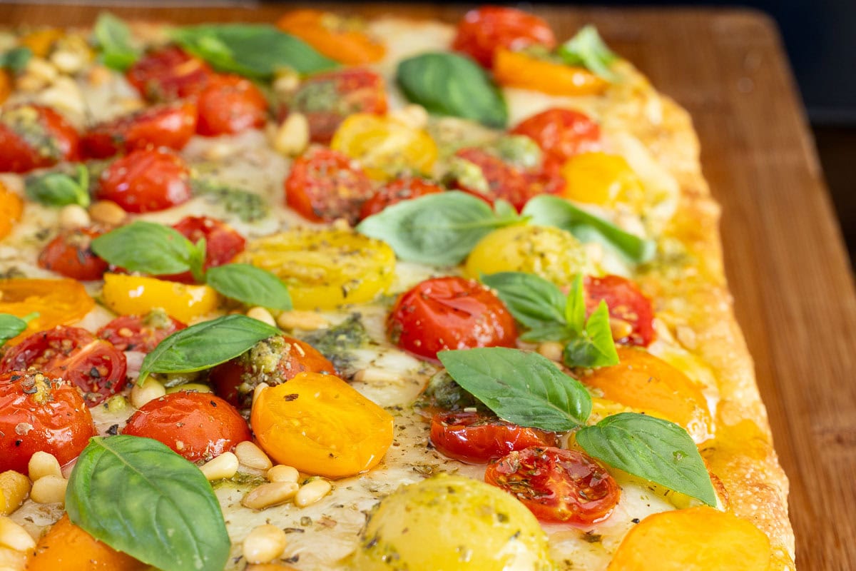 Close-up of Easy Sheet Pan Pizza Margherita with golden focaccia crust, roasted cherry tomatoes, pesto drizzle, pine nuts, and fresh basil