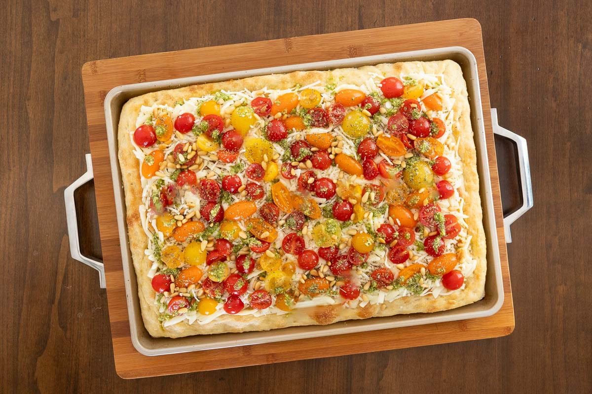 Unbaked Sheet Pan Pizza Margherita topped with mozzarella, salted cherry tomatoes, pesto drizzle, and pine nuts on focaccia crust