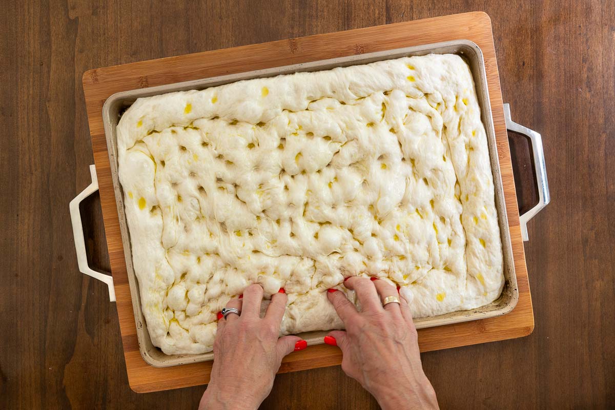 Hands dimpling risen focaccia dough with olive oil in a sheet pan to prepare the crust for Easy Sheet Pan Pizza Margherita