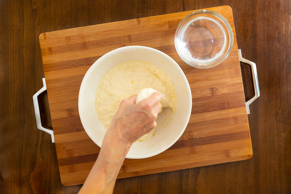 Hand folding risen focaccia dough in a white bowl to prepare the crust for Easy Sheet Pan Pizza Margherita