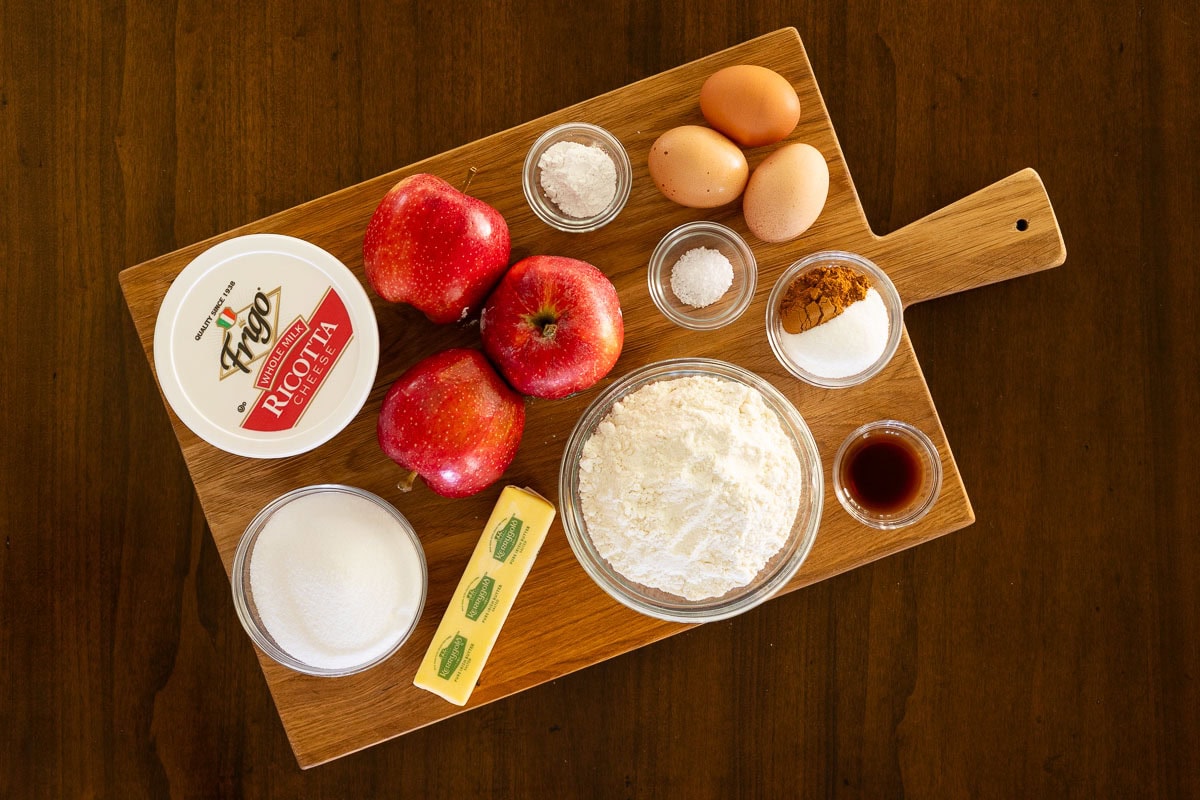 Ingredients for Italian Apple Ricotta Cake on a wooden board, including apples, ricotta cheese, eggs, flour, butter, sugar, cinnamon, and vanilla.