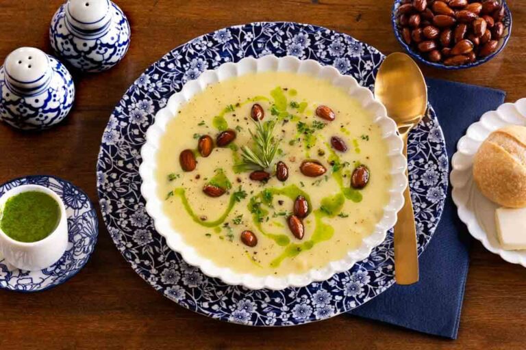 Bowl of creamy fall soup topped with roasted almonds, fresh rosemary, and herb oil, served with a crusty roll and butter on a wooden table.