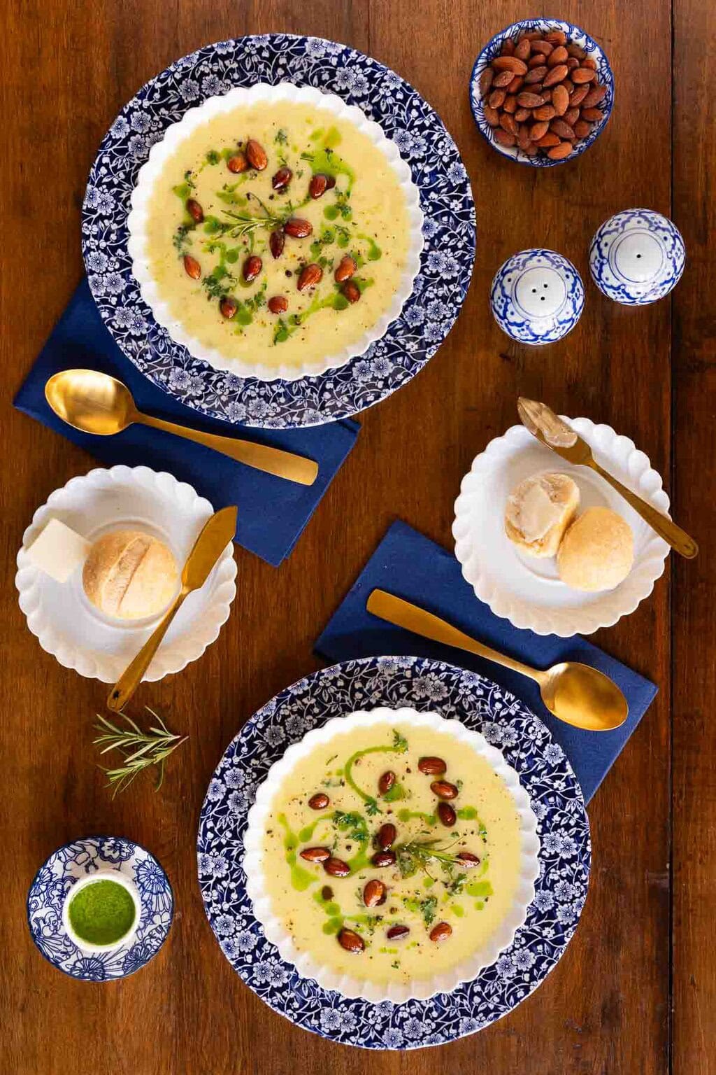 Two bowls of creamy fall soup garnished with almonds, fresh herbs, and a drizzle of green oil, served with crusty rolls on a wooden table.