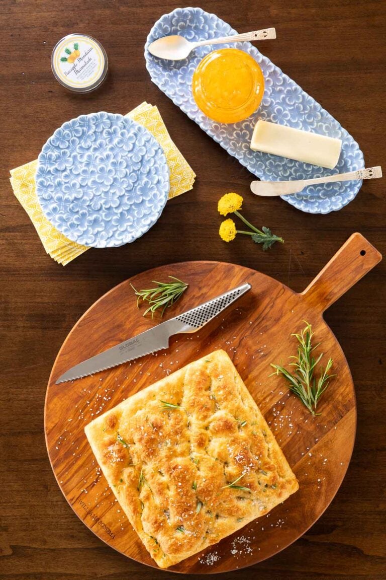 Golden square of rosemary focaccia bread on a wooden board with a serrated knife, fresh rosemary, butter, and marmalade on the side.