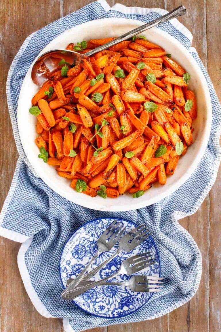 White serving bowl filled with roasted baby carrots, garnished with fresh herbs, with a spoon and a stack of blue-and-white plates on the side.