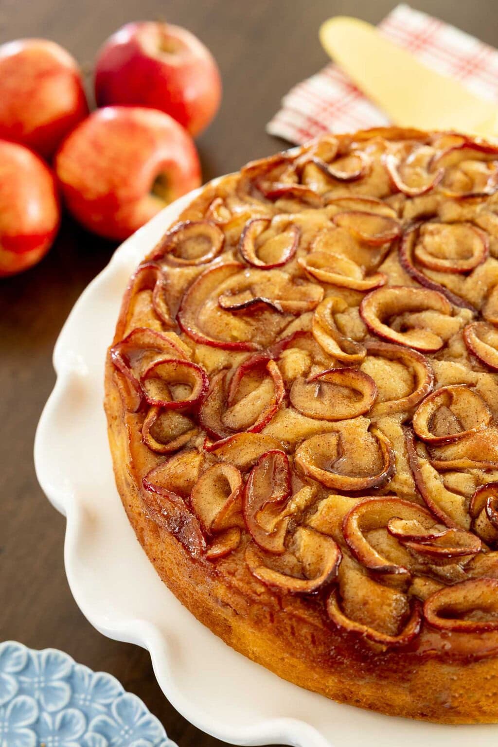 Close-up of Italian Apple Ricotta Cake topped with glossy cinnamon apple slices on a white scalloped cake stand, with fresh apples in the background.