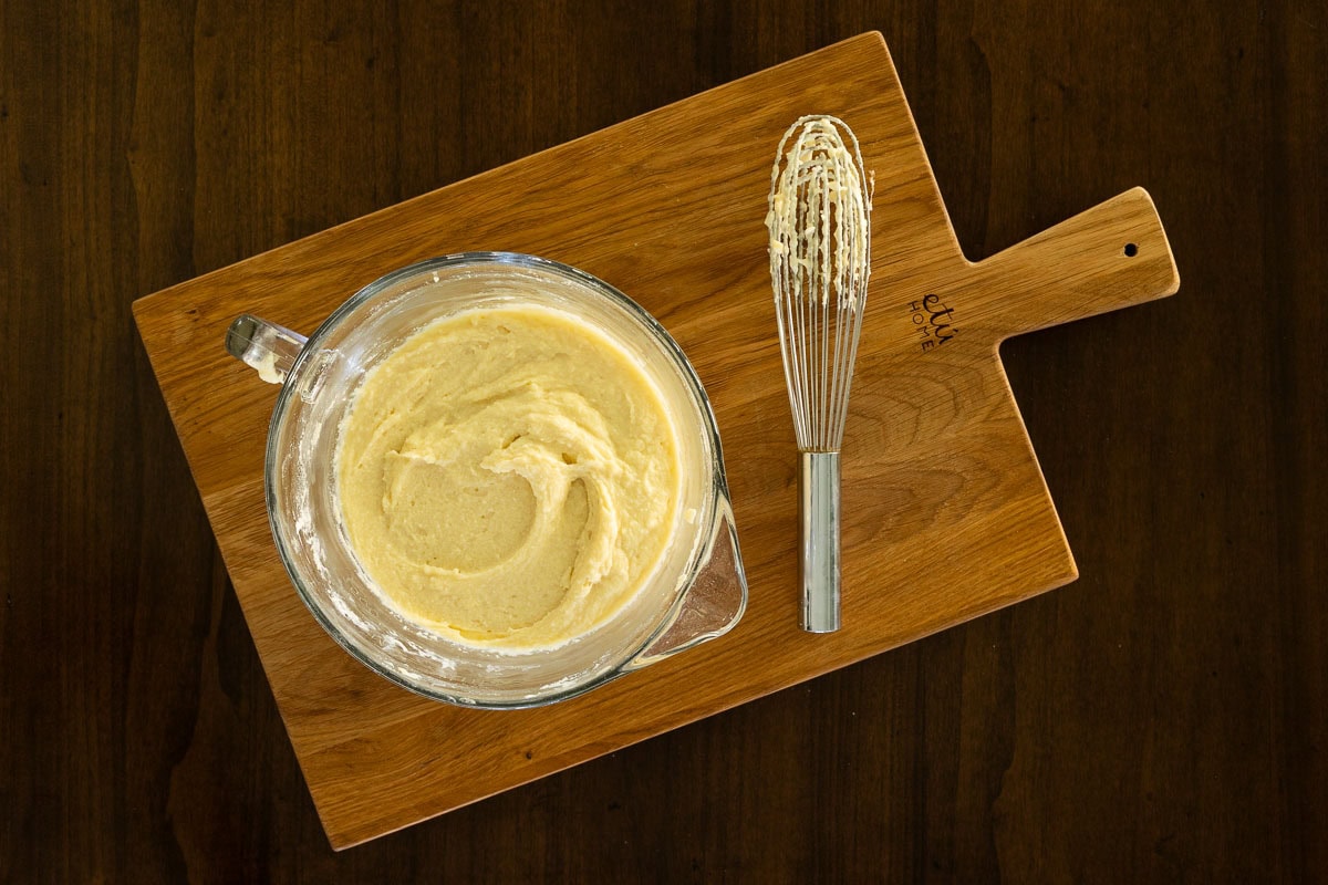 Glass mixing bowl of creamy ricotta cake batter with a whisk on a wooden board, showing the easy one-bowl preparation for Italian Apple Ricotta Cake