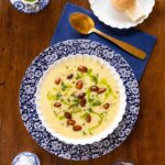 A bowl of creamy fall soup garnished with almonds, fresh herbs, and a drizzle of green oil, served with crusty rolls on a wooden table.
