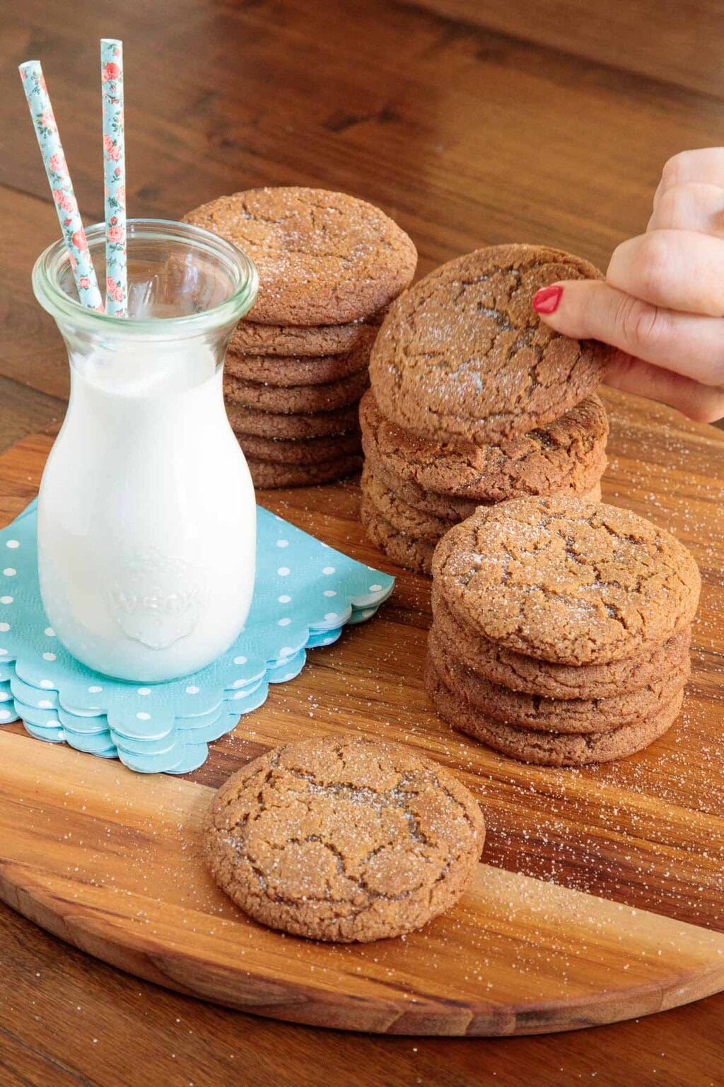 Hand picking up a Gingerdoodle Cookie from a stack beside a glass milk bottle with blue polka dot napkins and floral straws.