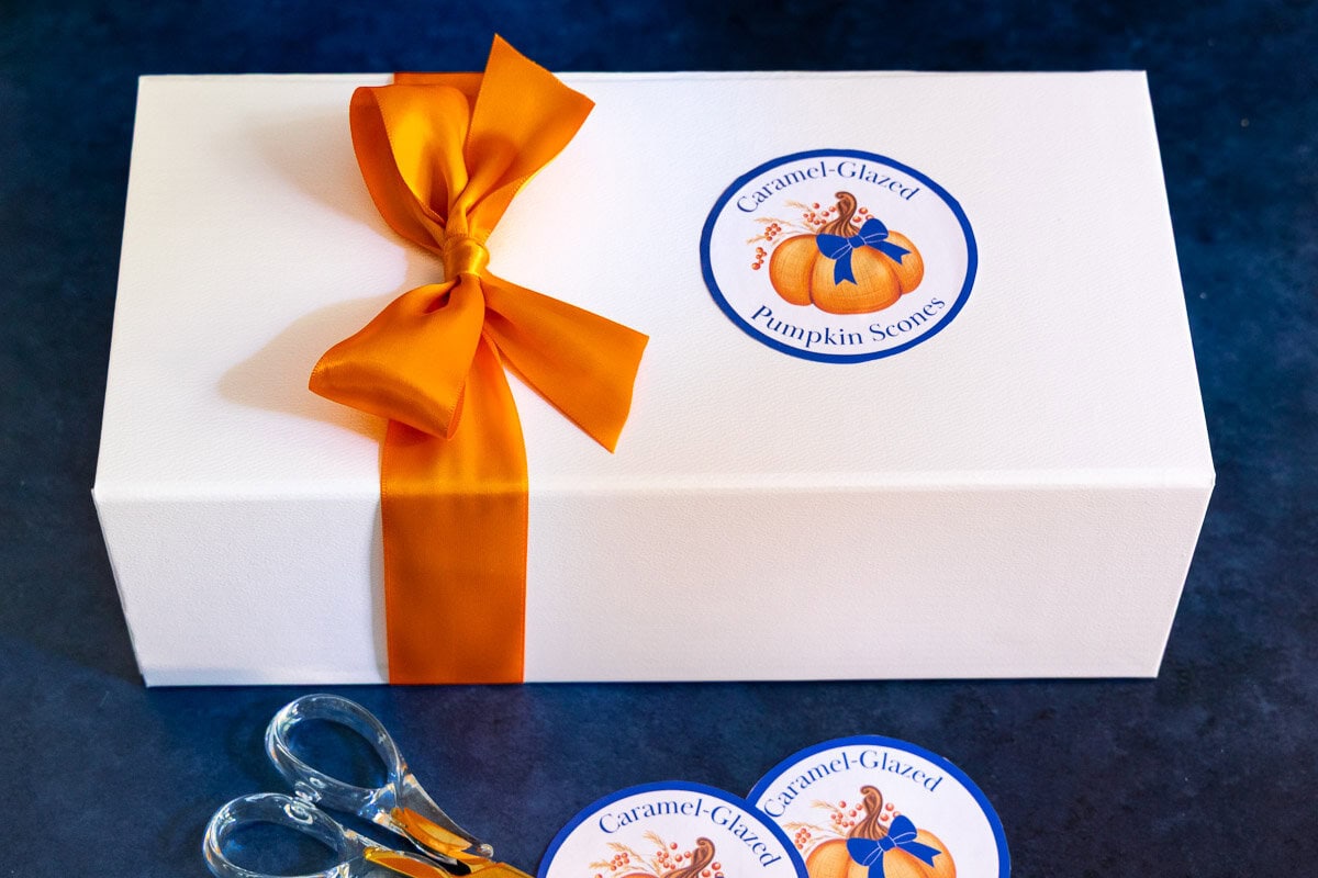 White gift box tied with an orange ribbon and labeled “Caramel-Glazed Pumpkin Scones,” with printable labels and scissors on a dark surface.