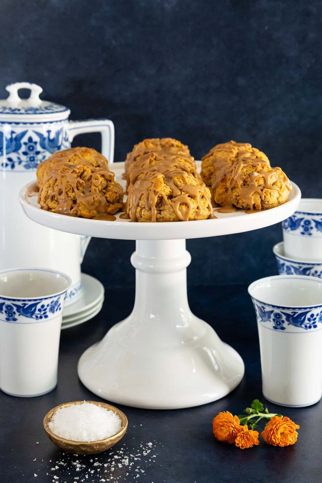 Pumpkin scones with salted caramel glaze on a white cake stand, styled with blue and white mugs, a bowl of flaky sea salt, and orange flowers.