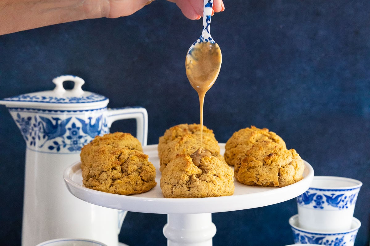 Hand drizzling salted caramel glaze over freshly baked pumpkin scones on a white cake stand with blue and white teacups in the background.