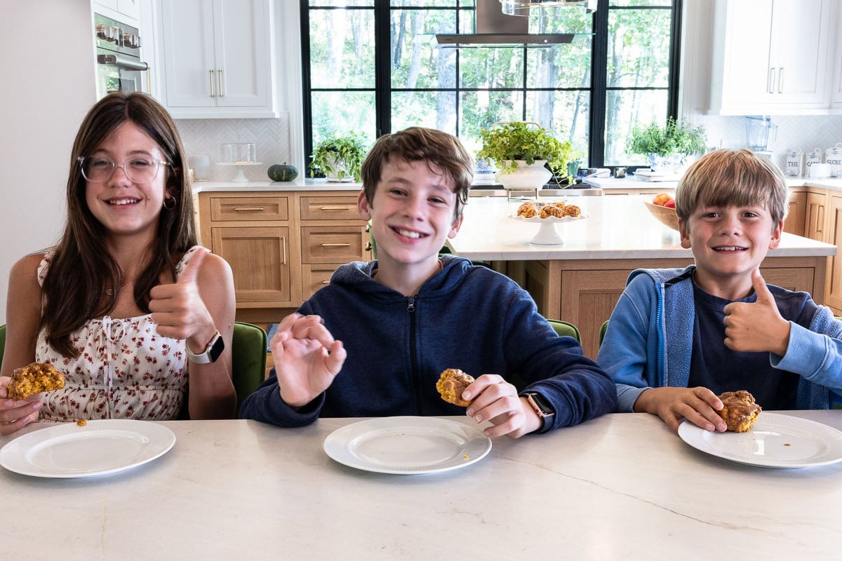 Three smiling kids giving thumbs up while enjoying pumpkin scones at a kitchen counter, with a cake stand of scones in the background.