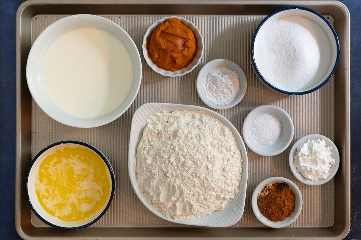Overhead view of ingredients for pumpkin scones on a baking tray, including flour, pumpkin puree, melted butter, cream, sugar, and spices.
