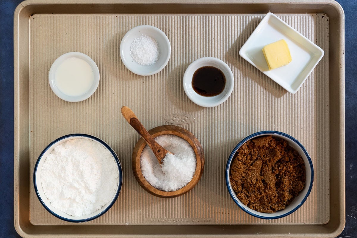 Overhead view of ingredients for salted caramel glaze on a baking tray, including butter, brown sugar, vanilla, cream, powdered sugar, and flaky salt.
