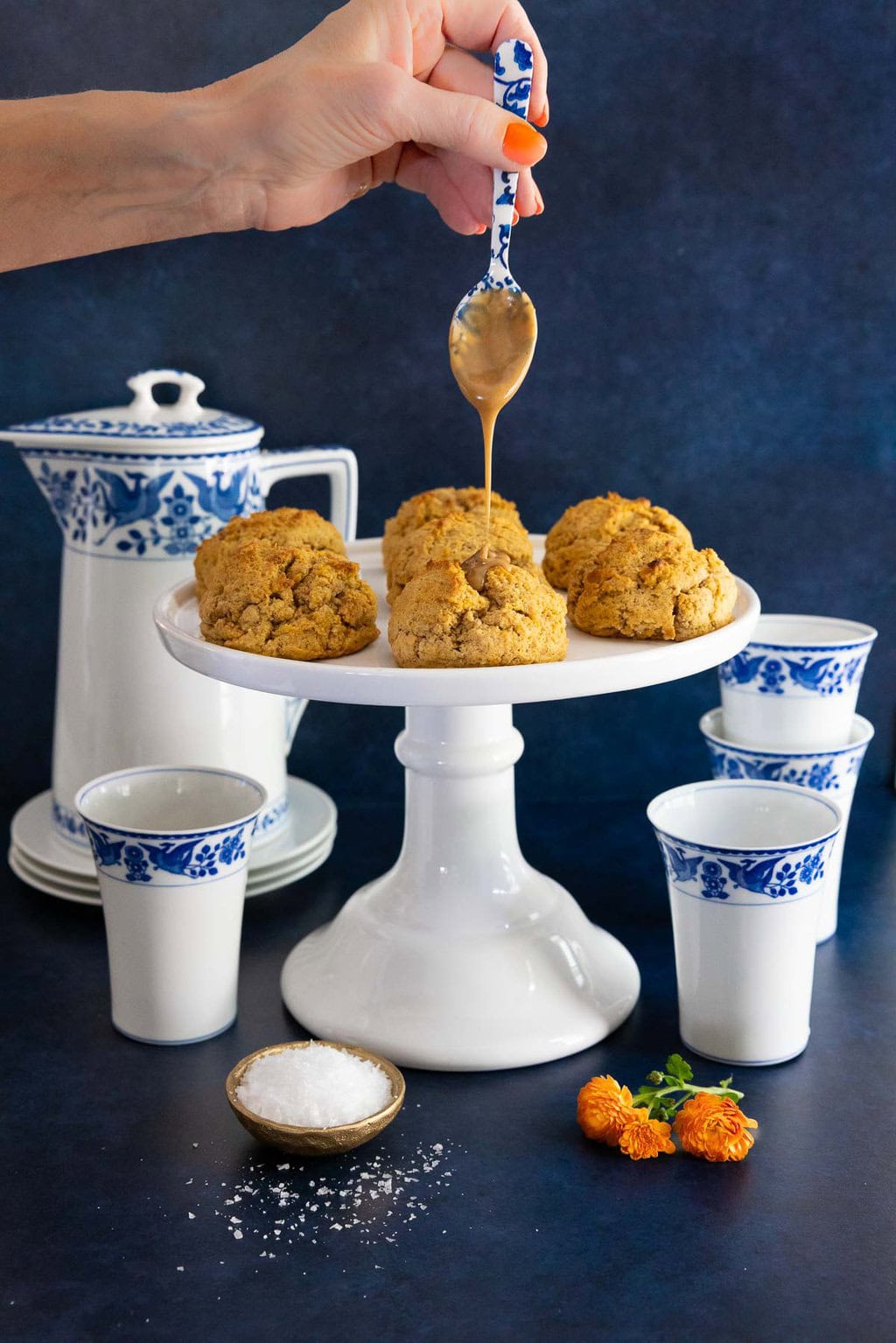 Hand drizzling salted caramel glaze over pumpkin scones on a white pedestal stand with blue and white cups and a bowl of flaky sea salt.