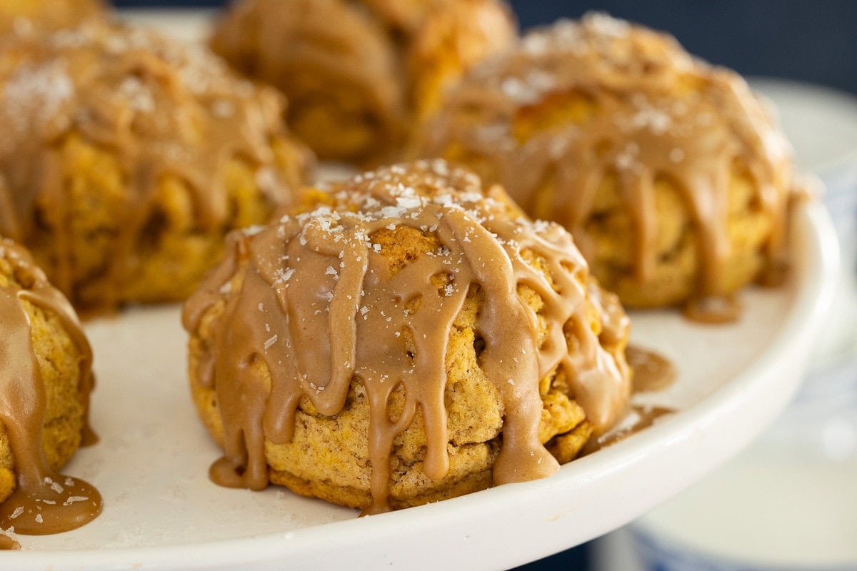 Close-up of pumpkin scone drizzled with salted caramel glaze and sprinkled with flaky sea salt on a white cake stand.