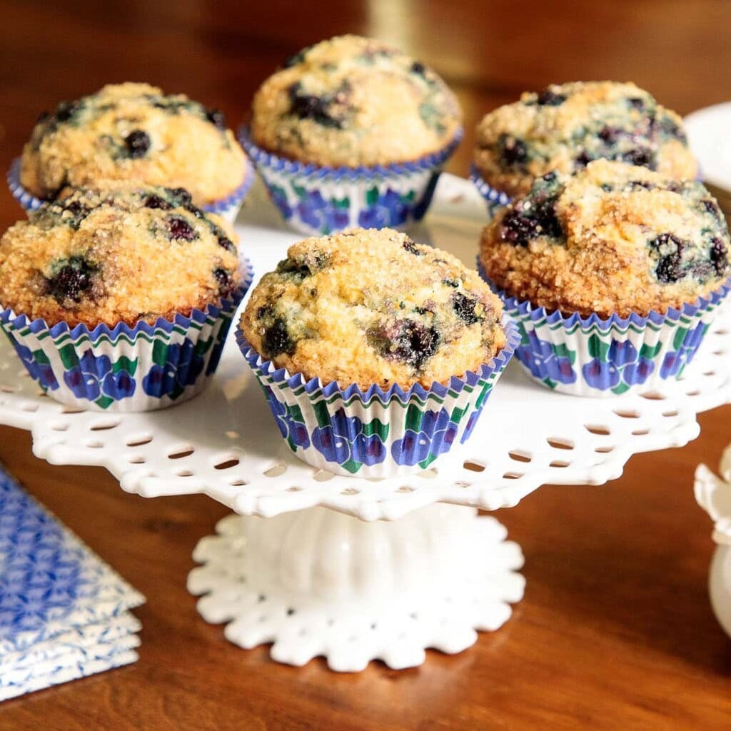 Six bakery-style blueberry muffins on a white cake plate.